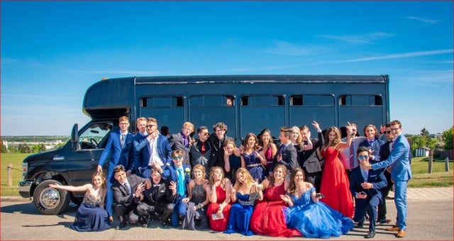 High school graduates in suits and gowns take group photo in front of black party bus with blue skies in background