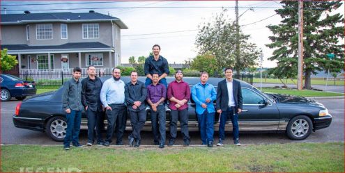 8 guys standing in front of and one man sitting on top of a Black Lincoln Stretch Limousine