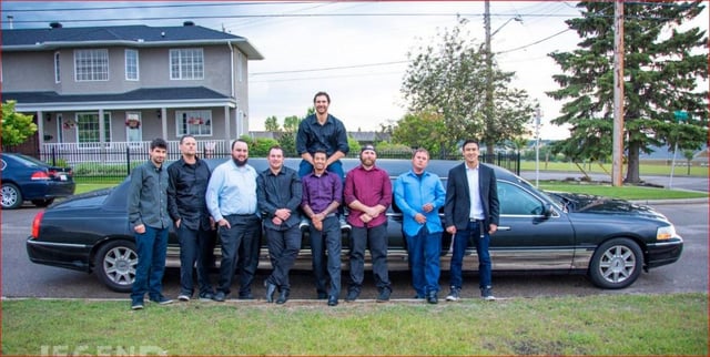 8 men standing in front of black stretch limousine with one man sitting on top