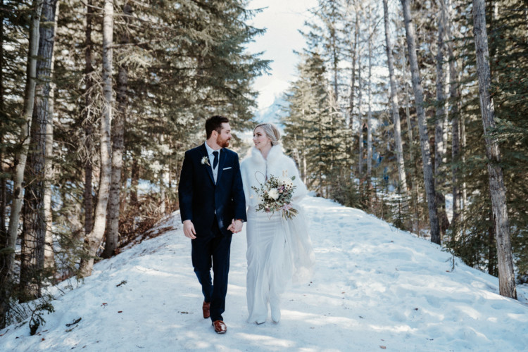 Photo of bride and groom walking on snowy trail between trees