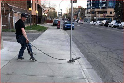 Man standing on sidewalk with a chain from his ankle to a metal pole with a metal male body part attached for stag party