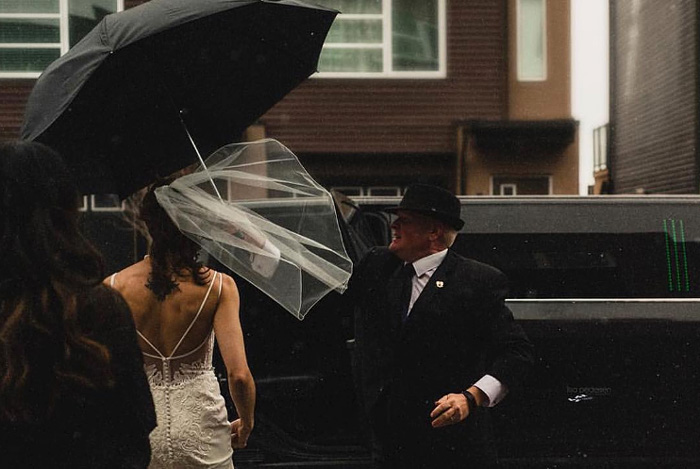 Limo driver holding black umbrella over bride in white gown while she enters black limousine